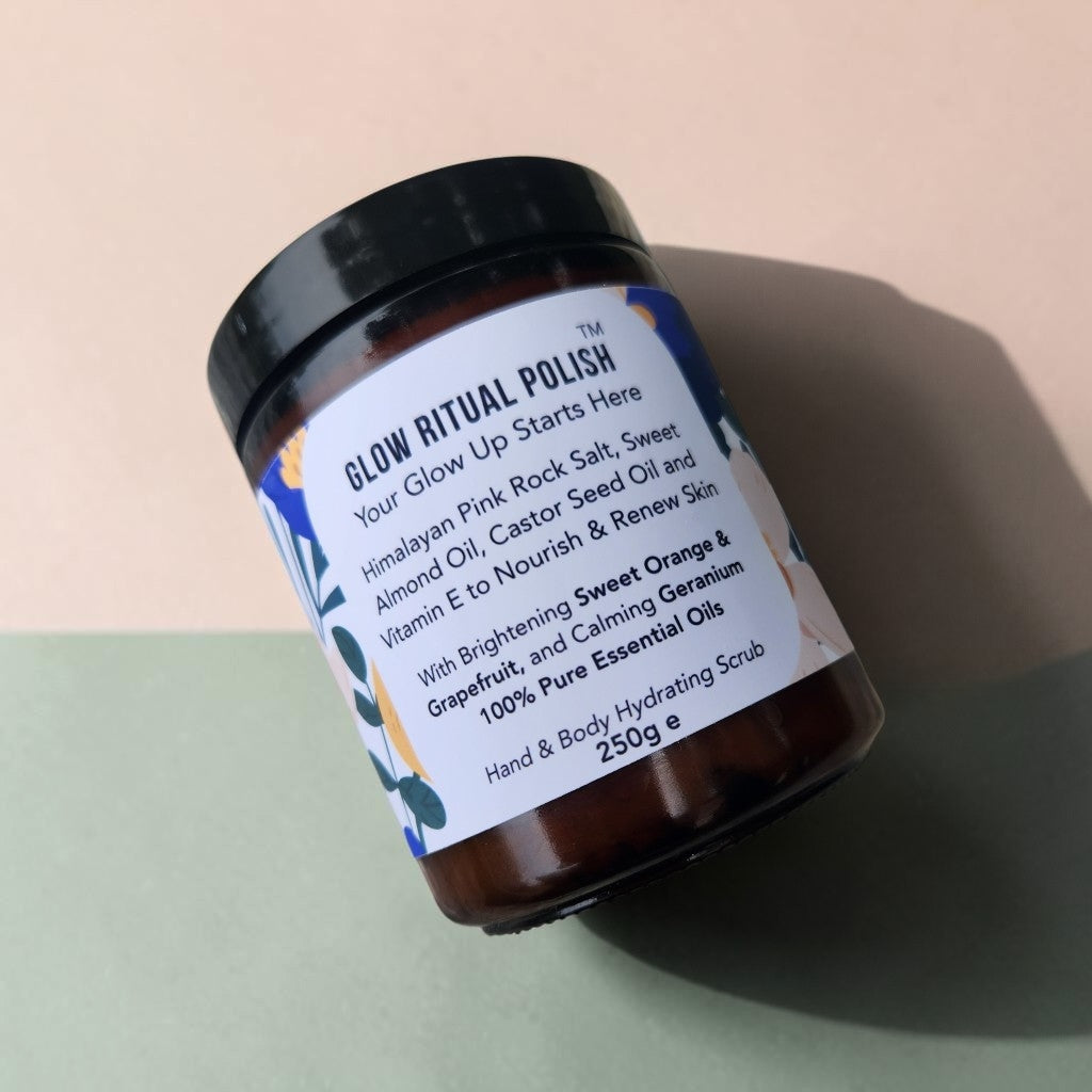 A jar of Glow Ritual Polish hand and body hydrating scrub, featuring a black lid and a white label with blue floral accents, set against a split beige and sage green background.