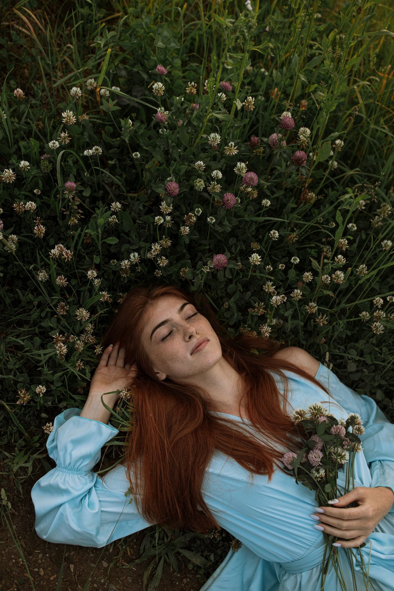 A woman with long red hair lies on her back in a field of wildflowers, wearing a light blue off-the-shoulder dress and holding a small bouquet of flowers.