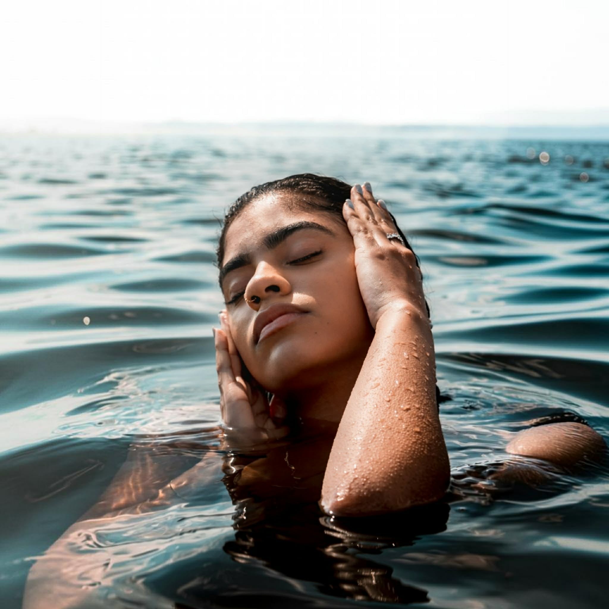 A woman with her eyes closed floats in dark blue water, holding her face with both hands. Her skin is glistening with water droplets, and the ocean surface surrounds her under a bright, hazy sky.