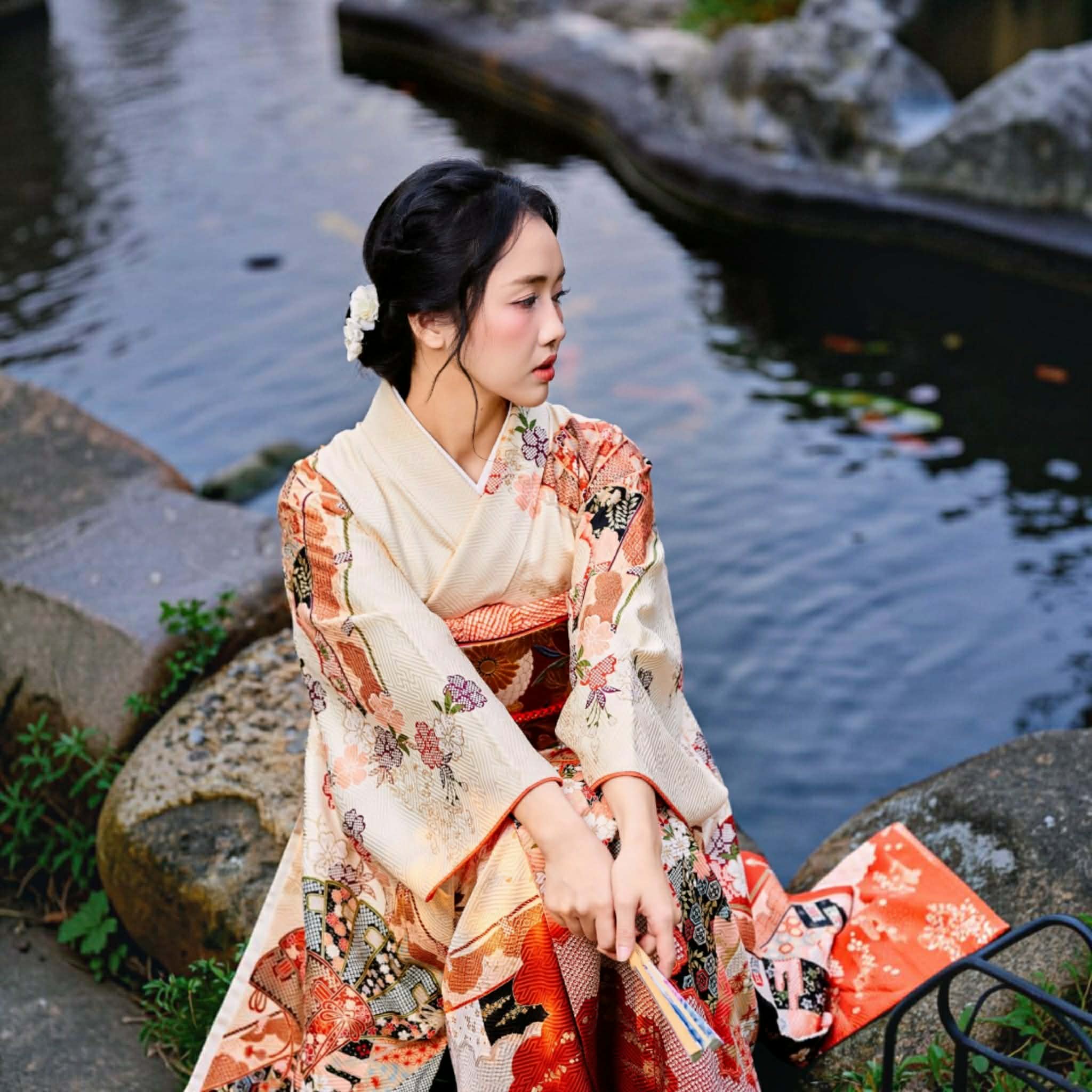 A woman wearing a traditional Japanese kimono sits on a stone beside a pond, holding a folding fan. The kimono has a cream base with orange, red, and floral patterns.
