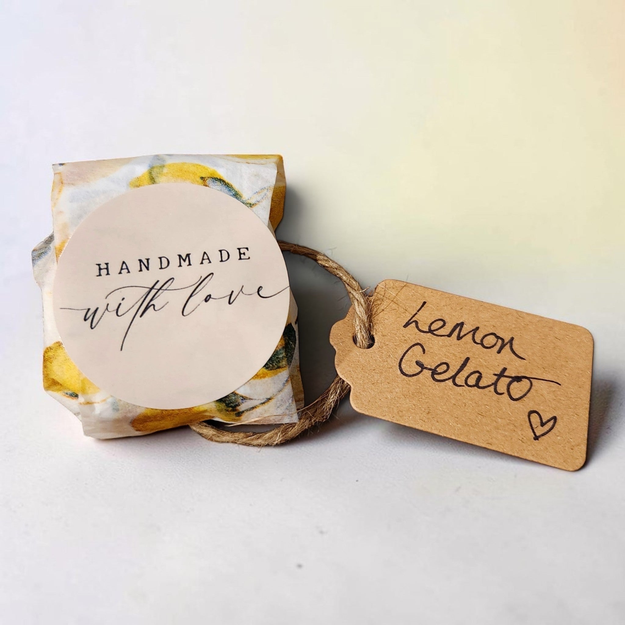 A small wrapped gift box with a round cream paper label reading 'HANDMADE with love' and a kraft paper hang tag tied with twine that says 'Lemon Gelato' in black script, photographed on a light neutral background.