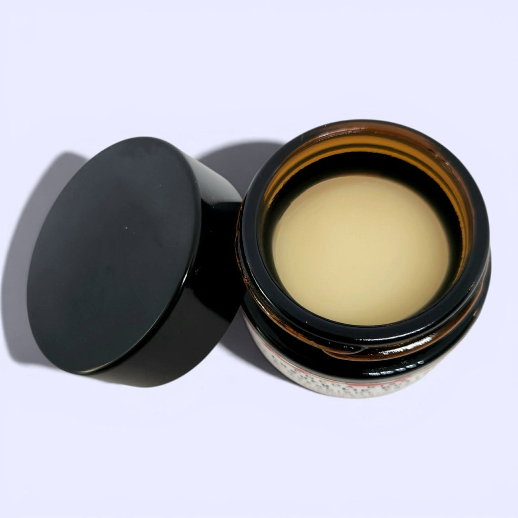 An open amber glass jar containing a pale yellow cream or balm, with its black lid resting beside it on a light lavender background.