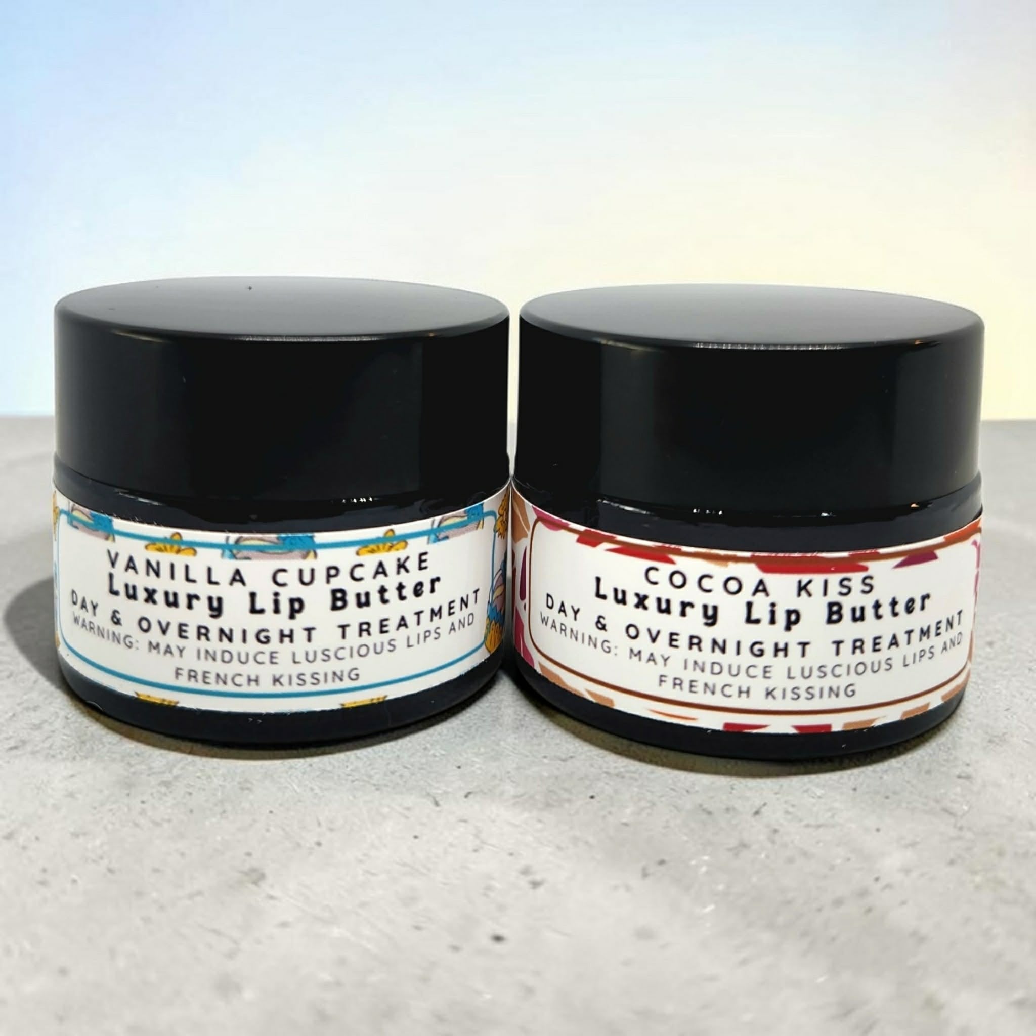 Two small amber jars of luxury lip butter on a light gray surface against a pale blue background. The left jar is labeled Vanilla Cupcake and the right jar is labeled Cocoa Kiss, both with white labels and black text.