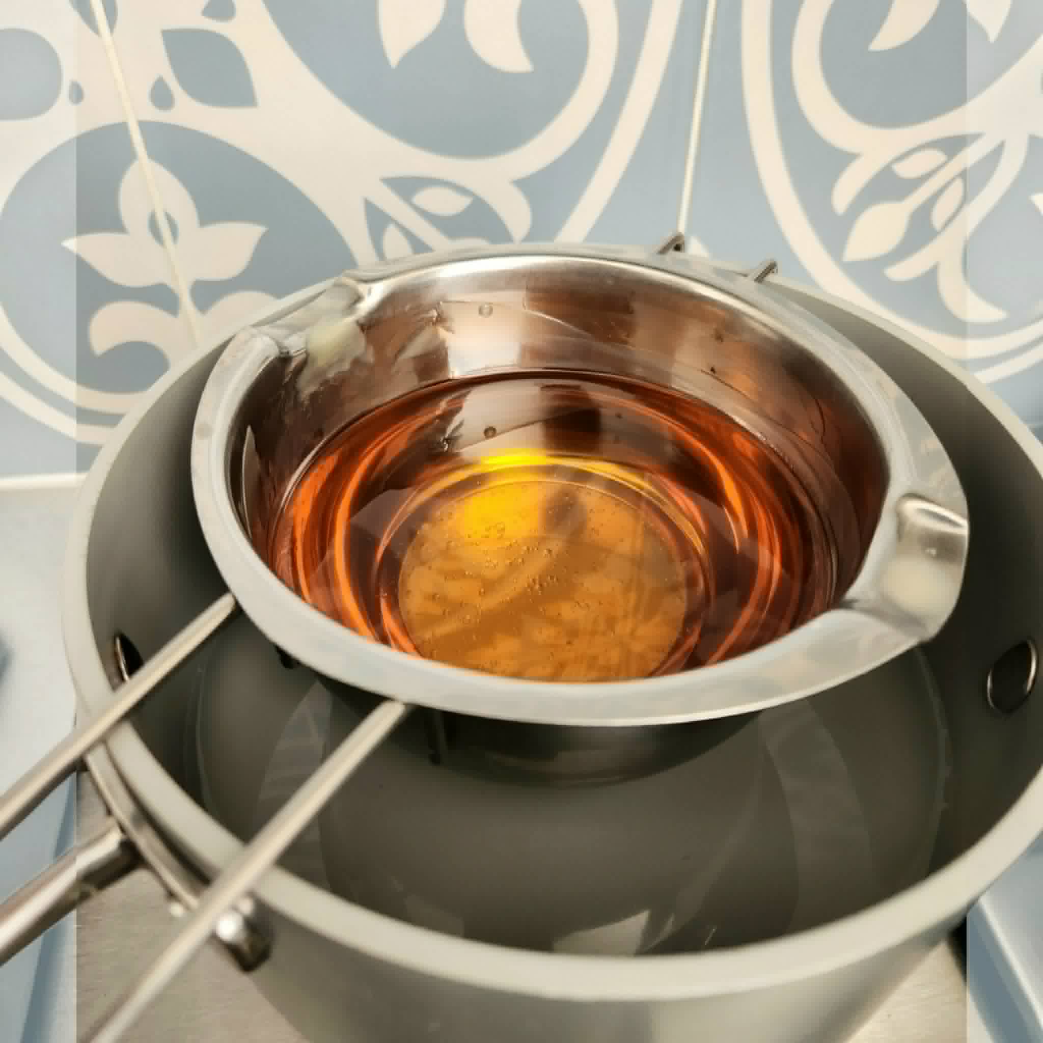 A stainless steel double-boiler setup with a smaller pot inside a larger pot, holding amber-colored liquid in a glass or clear container, photographed against a blue and white patterned background.