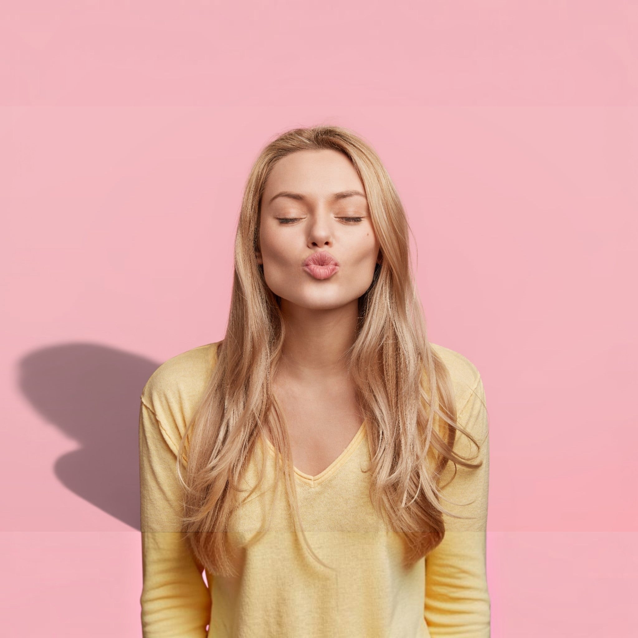 A blonde woman with her eyes closed and puckering her lips, wearing a pale yellow long-sleeve V-neck top against a solid pink background.