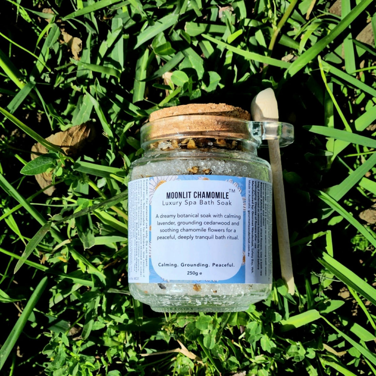 A glass jar of Moonlit Chamomile Luxury Spa Bath Soaks sits on a bed of green grass. The clear jar has a natural cork lid, a white label with blue and orange text, and a small wooden spoon rests on the right side.