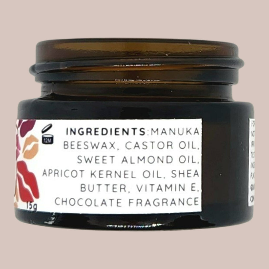 A dark amber glass jar of skincare cream with a white label detailing ingredients such as manuka beeswax and castor oil, set against a solid beige background.