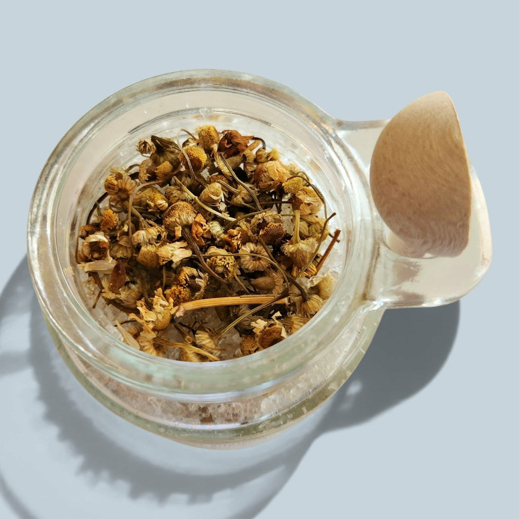 An overhead close-up of a clear glass jar filled with dried chamomile flowers and a small wooden spoon, set against a light blue-gray background.