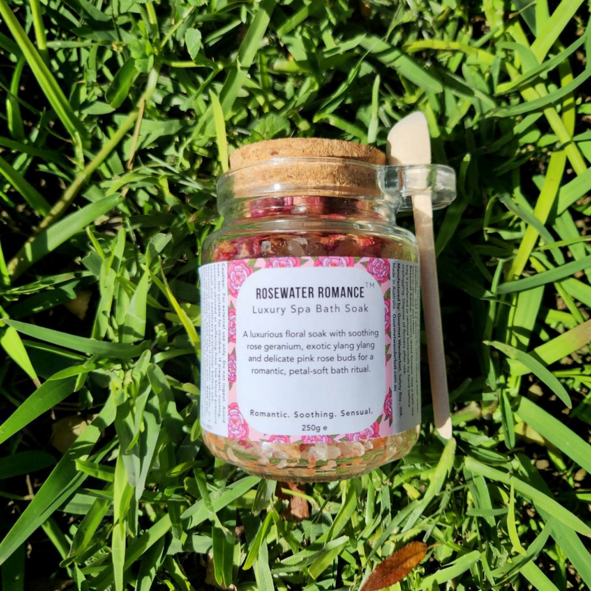 A glass jar of Rosewater Romance Luxury Spa Bath Soak with a cork lid and a wooden spoon, placed on a bed of green grass in natural sunlight.