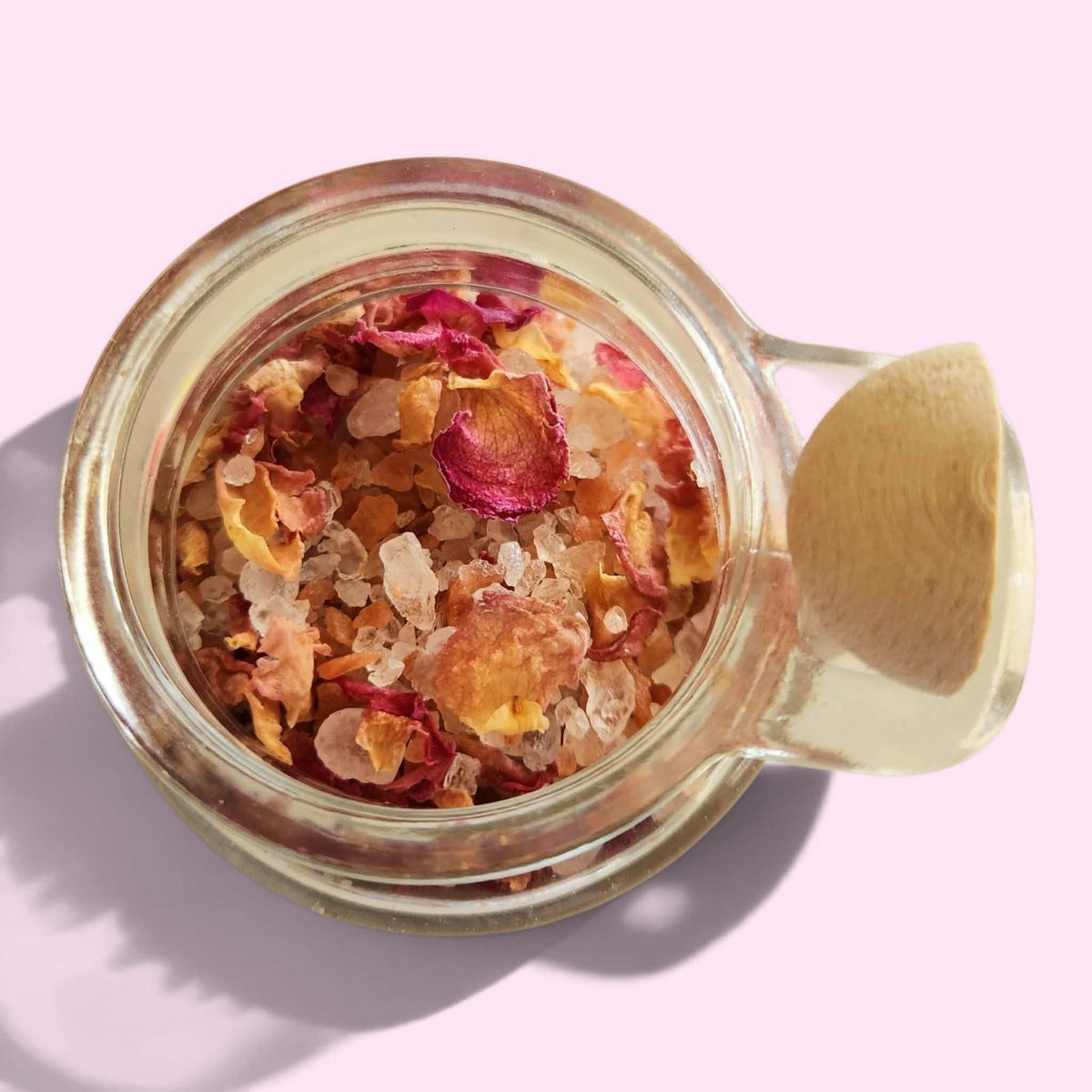 A top-down view of a clear glass jar filled with dried flower petals and coarse salt crystals, with a small wooden spoon resting inside, set against a solid light pink background.