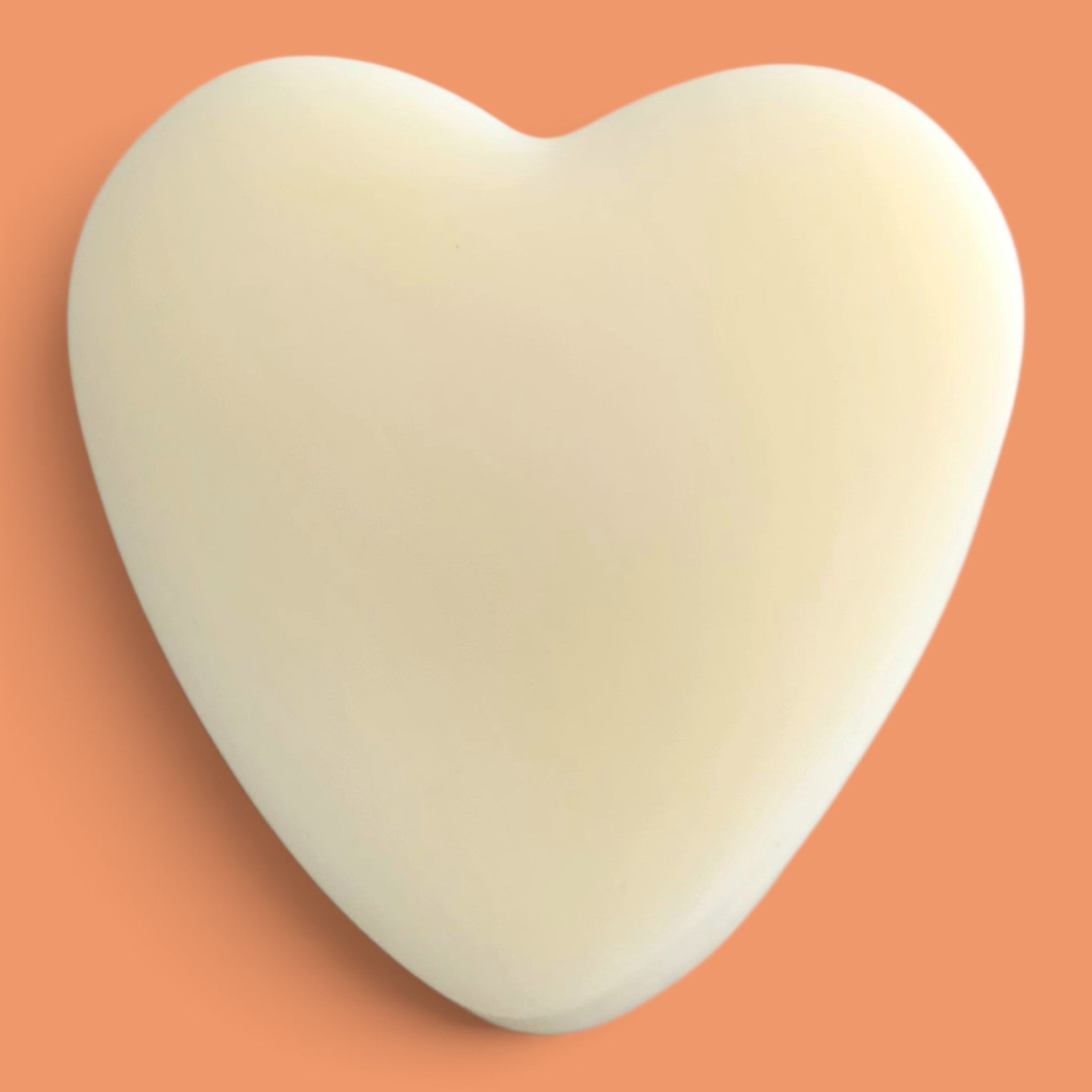A smooth, heart-shaped cream-colored object, resembling a silicone bath bomb or soap bar, centered on a solid peach-orange background.