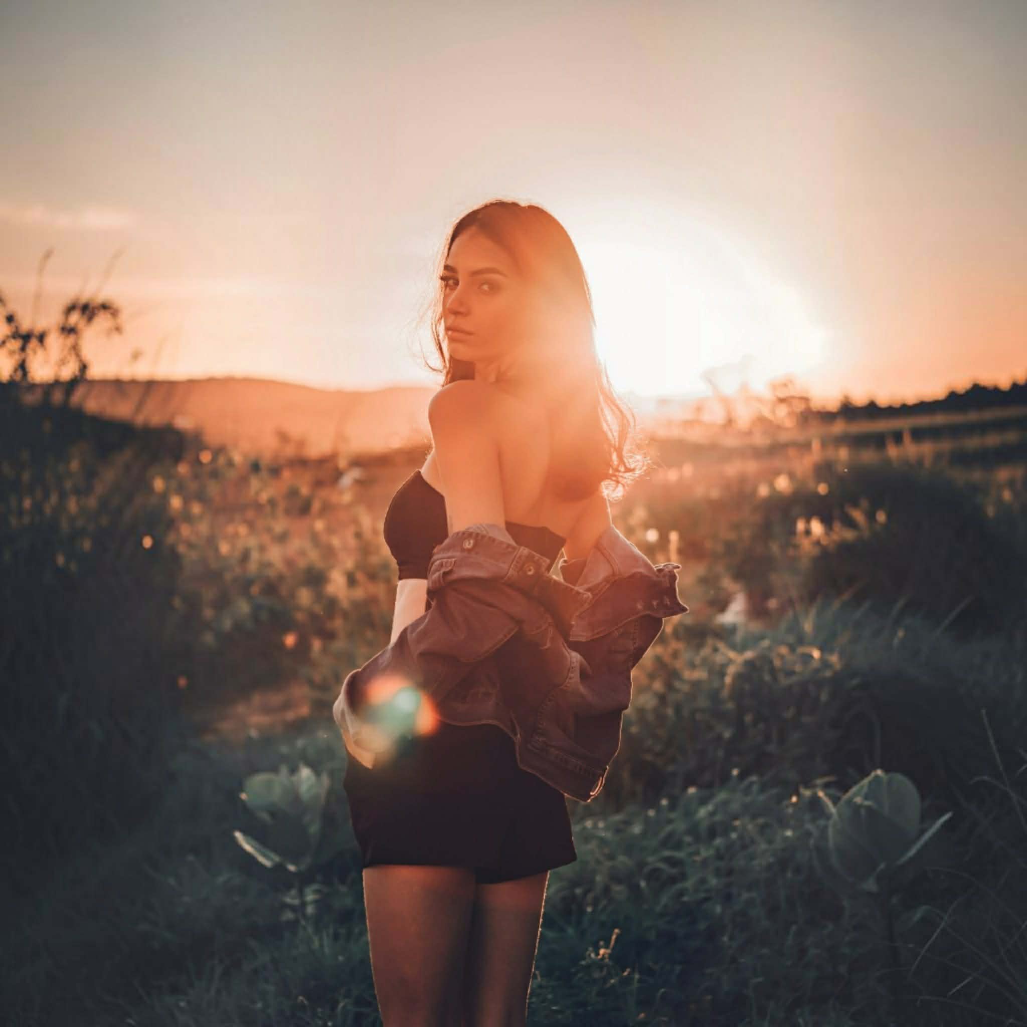 A woman stands outdoors in a field at sunset, wearing a black strapless top and a short black skirt, with a dark draped jacket or shawl around her shoulders. The sun is low in the sky, creating a bright flare behind her.