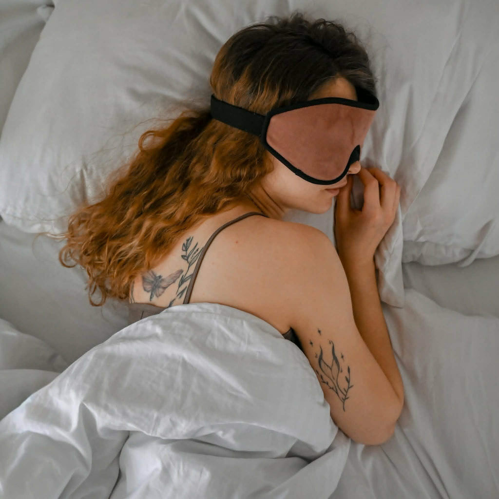 Person wearing a sleep mask lying in bed with white bedding