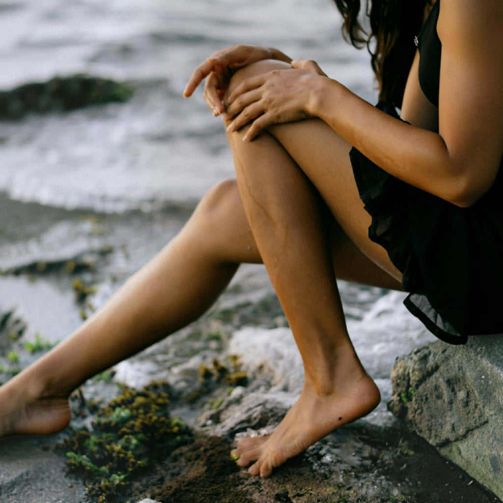 A woman sitting barefoot on rocks by the ocean, wearing a black sleeveless dress. Her legs and hands are visible as she rests her arms on her knees, with waves and wet stones in the background.